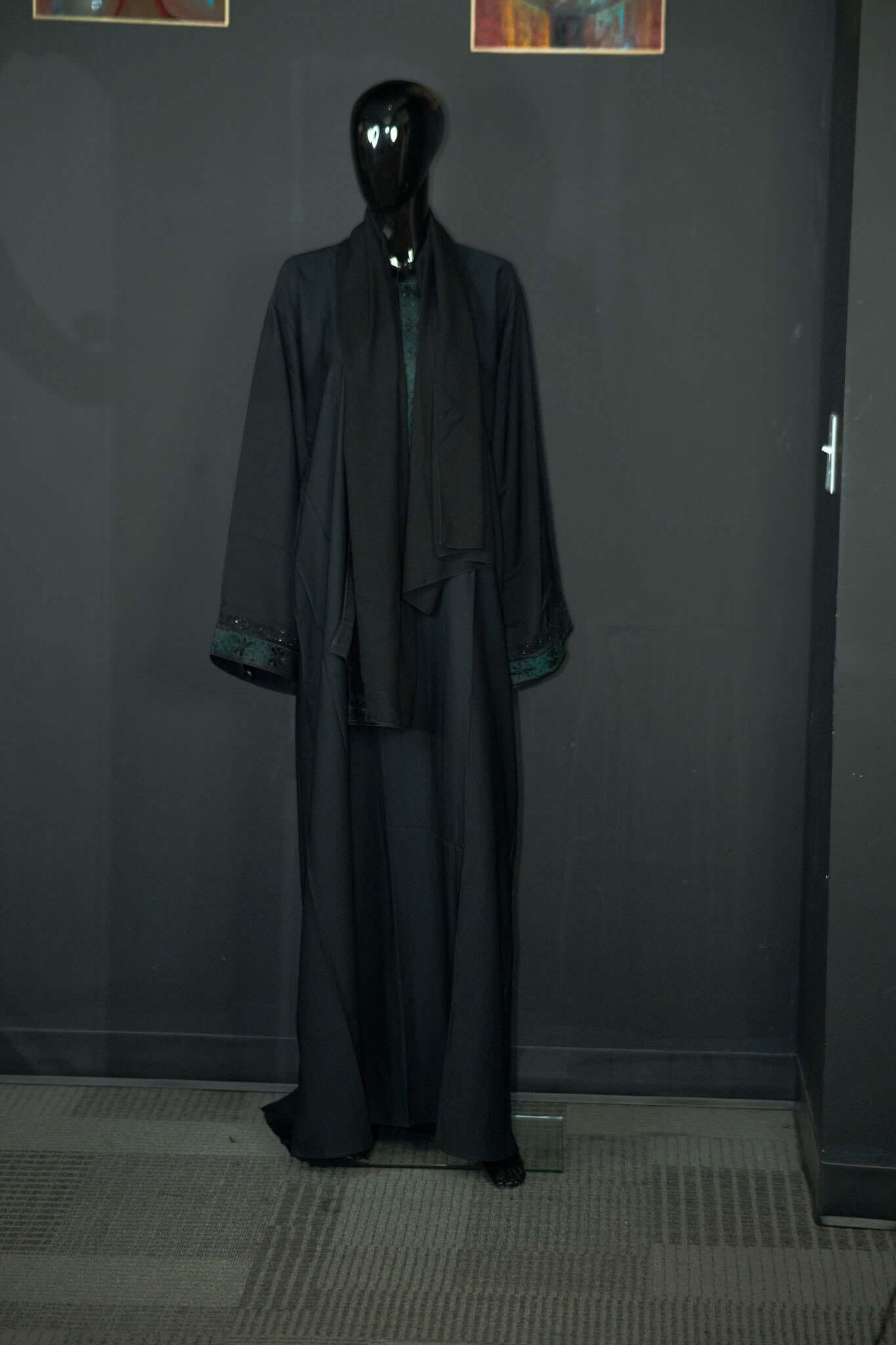 Black abaya with intricate designs by Oumy Beauty Studio displayed on a mannequin in a dark room.